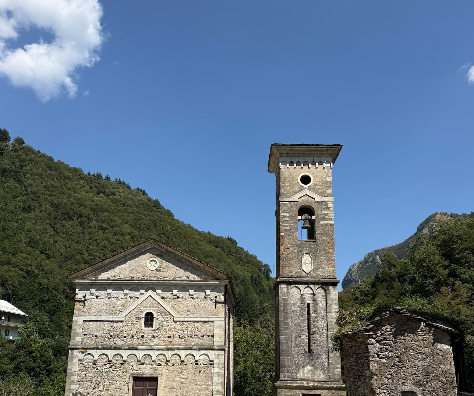 A stone church and a tall separate campanile against a wooded mountainside